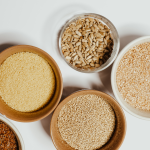 A top-down view of various whole grains and seeds in bowls, including sunflower seeds, quinoa, cousc