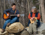 Jess Martin playing a guitar sitting on a stone wall next to Kim Hoff who is holding a book.