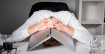 An office worker hunched over their desk in despair with their laptop folded over their head.