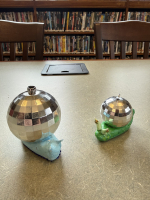 Two snails made out of clay with disco balls for shells on a table in front of a bookshelf