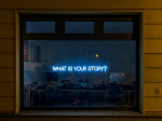 A neon light in a window reads "What is your story?"