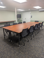 Large room with light walls and dark carpet, and a group of wooden tables and black chairs arranged 