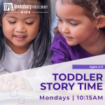 Two toddlers are smiling and sitting next to each other. One is reading a book and the other is look