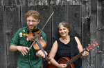 A white man holds a fiddle next to a white woman holding a guitar. Both are smiling. 