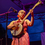 A woman with glasses in a red dress holds a banjo in front of a microphone.