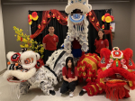 Three people in red shirts pose next to traditional lion dance equipment against a black backdrop wi