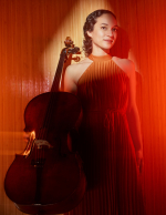 a woman in an orange dress stands with a cello against an orange backdrop