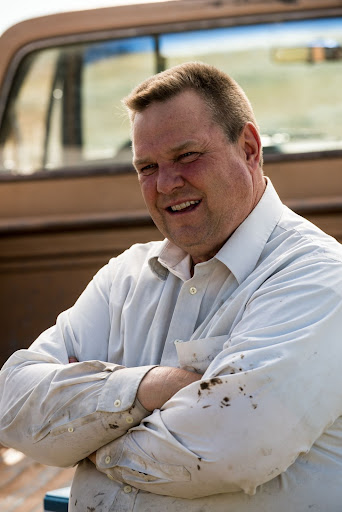 Sen. Jon Tester with arms crossed in dirt-stained t-shirt standing in front of pickup truck