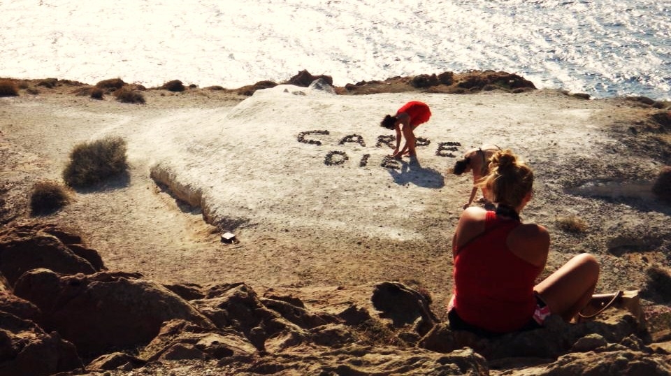 [Is there any life out there? In the sparkling sun of the ocean...] At the southernmost tip of Santorini, I watch two girls arrange rocks into the words 'Carpe Diem'. A couple minutes later and the words would have already been fully formed, but the moment is perfect; I am capturing the action. A girl sits above them, directing their movements, and the sun sparkles down over us, rippling fragments of light bobbing on the ocean's enthusiasm, urging us to seize the day.