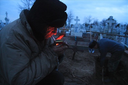 Pavel Hancearencu, the town's gravediggers in Sulina, lights a cigarette while his nephew, Augustin Hriscenco, takes turn on digging the grave for the next day funeral, on Christmas Eve. Sulina, Danube Delta, Romania.