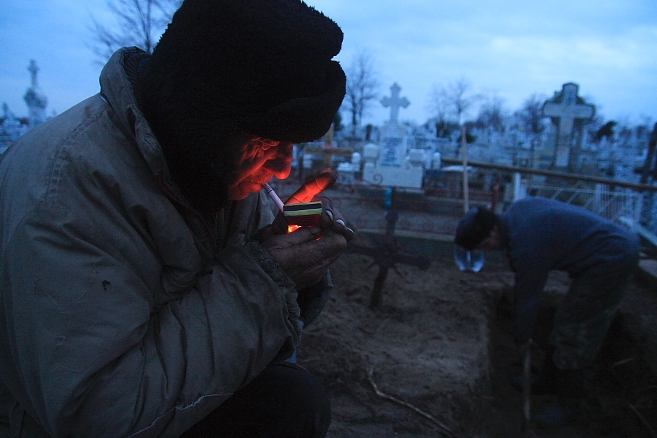 Pavel Hancearencu, the town's gravediggers in Sulina, lights a cigarette while his nephew, Augustin Hriscenco, takes turn on digging the grave for the next day funeral, on Christmas Eve. Sulina, Danube Delta, Romania.