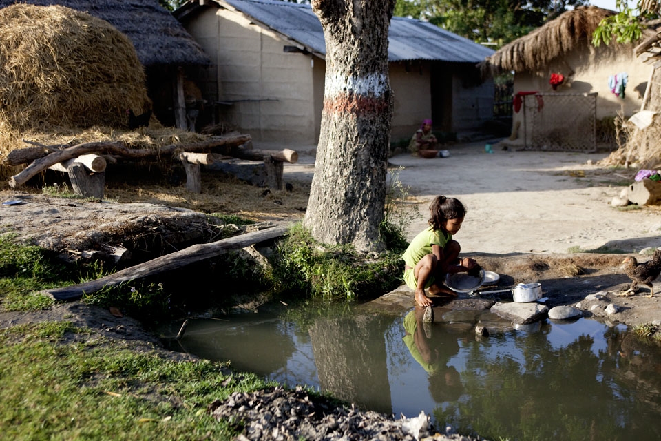 Young girl washing dishes in polluted river while her mother is cooking behind.