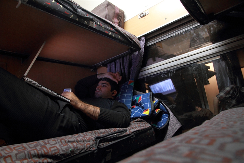 A man is travelling on a ‘6-bed first class’ and lying down on a ‘second floor’ bed. There are 3 rows of beds which can be folded and fall back on the wall. Once folded, the first row becomes the bottom seat, and the second row the back seat. Train and tracks are often old and slow. Many prefer to pay a bit more to be able to sleep during the long travels.