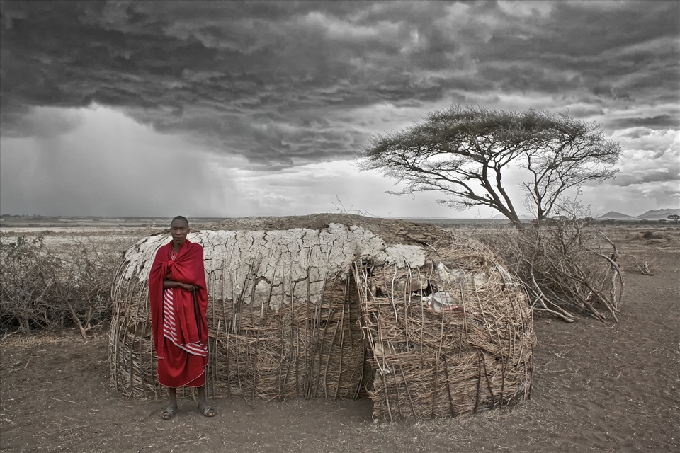 A Masai Warrior stands proud outside his home as a storm gathers over the plains