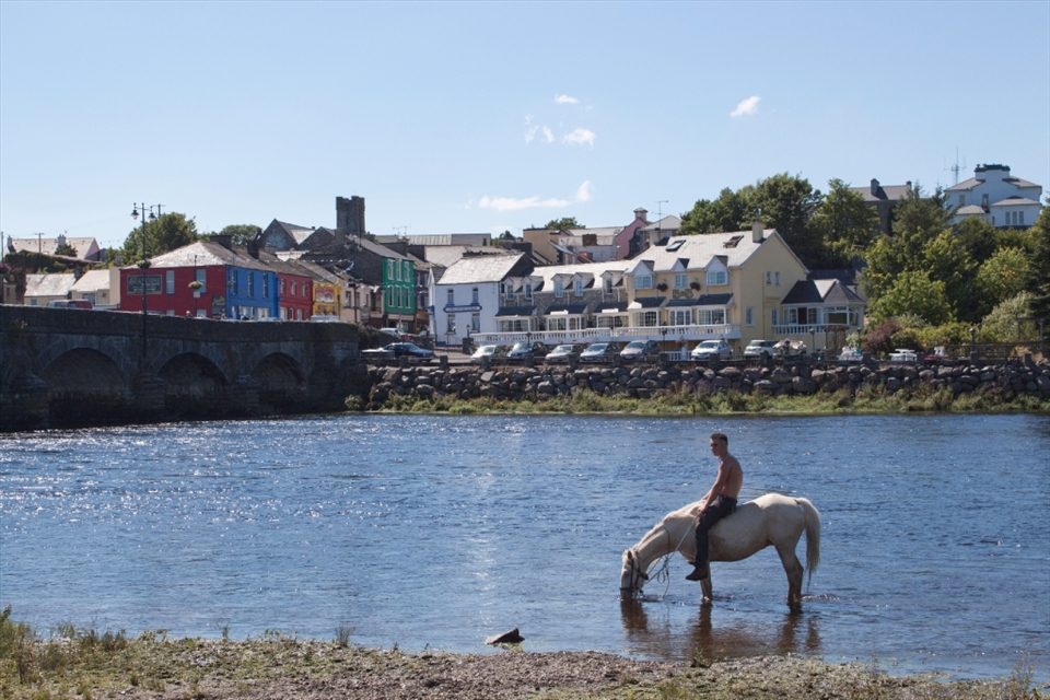 The early morning horse fair ended, a Gypsy boy rides his horse bareback into the River Laune to escape the heat and dust while the colorful town of Killorglin sits in the background.