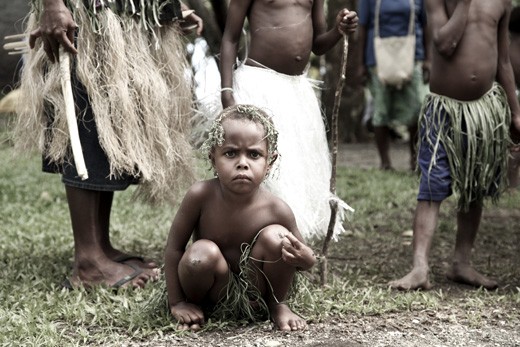 A child wearing a grass skirt squats beside a path. To make money for their families, they are sent out to sing and dance for the tourists; something most children do for fun or as part of imaginary games, but never for their livelihood. 