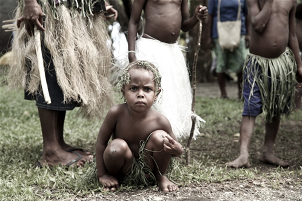 A child wearing a grass skirt squats beside a path. To make money for their families, they are sent out to sing and dance for the tourists; something most children do for fun or as part of imaginary games, but never for their livelihood. 