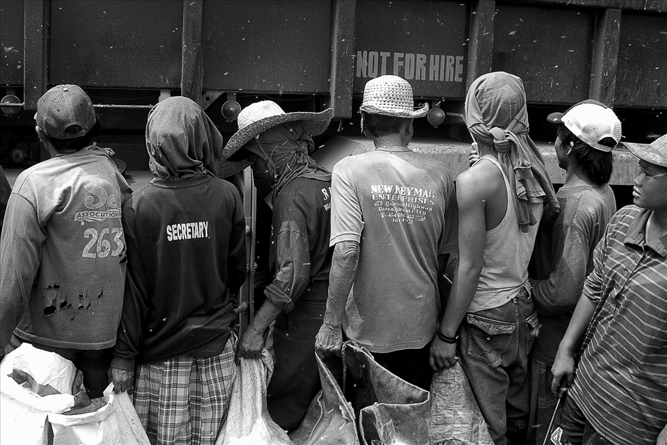 Scavengers line up beside a dump truck which is about to empty its contents. Competition is intense to collect the best items in the tons of waste disposed here every day. Conditions are dangerous and very unsanitary. The decomposing biomass attracts many flies. 
