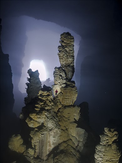 Our expedition leader Watson climbs a huge stalagmite to help photograph scale. From this particular vantage point, the 'Witches Hat' formation can be seen in the back ground, a unique silhouette, due to the glow from the first doline.