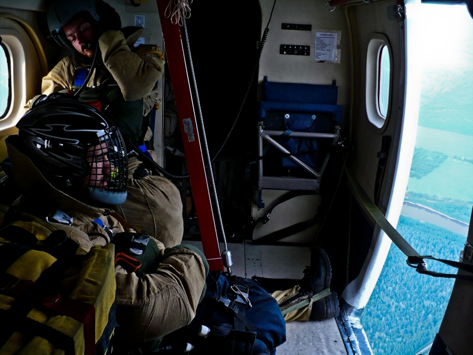 Two Smokejumpers, en-route to a fire, sit patiently and watch the world go by through the opening they will soon depart through.
The use of fixed-wing aircraft and parachutes allows for crews to be placed on fires in remote locations far more quickly than other methods; allowing for fires to be extinguished quickly and before they grow too rapidly.