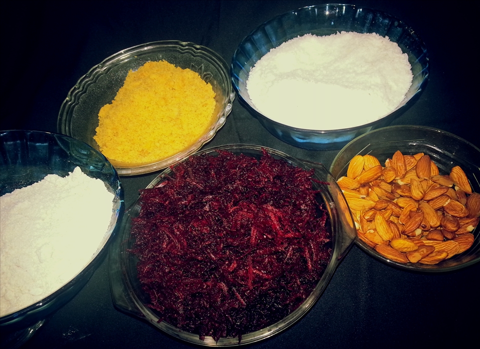 the ingredients of the dish - center maroon color is - grated beetroot, on the right to beetroot is almond, behind almonds there is a bowl of coconut shavings,beside it lies jaggery powder and on to the left of jaggery lies blended/powdered oats