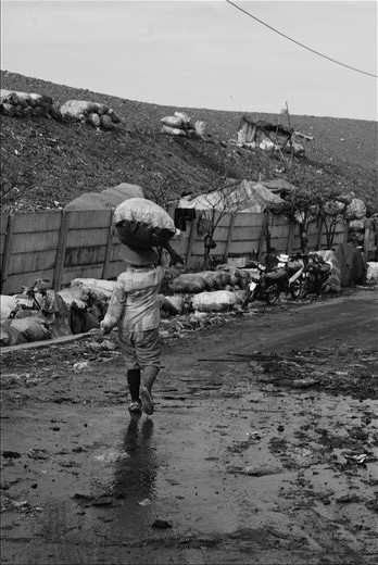 Carrying a bag full of waste on this old lady’s head accompanied by her own shade reflected in puddle and slippery road is very unique image; with the overloaded weight, she can walk calmly enjoying every step knowing this bag would be pondered in weighing location which located in the entrance and needs more than one hour walk from her location at that time.  Their struggle for life, dedication to their families, willingness to live in that slum area for funding the living, steadfast hardworking effort, and commitment in each family member to strive together facing the life problems warn the society to consider poverty which is also able to haunt them and also confront the sumptuous and high-priced city’s life style contrasting with this people life. Will the society or government provide them with better 