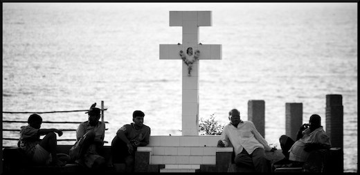 A cross stands tall against the mighty ocean as the locals chill around it.