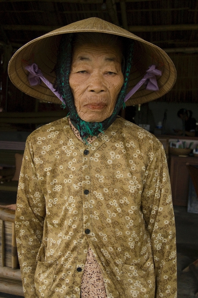 This almost lifeless woman keeps up tradition wearing a conical hat, matching her strong yellowing skin and dowdy shirt. She looks almost like a chiseled sculpture. The tired look perhaps one of her last!
