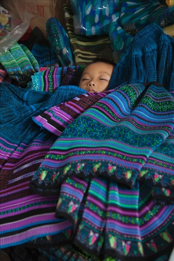 Fluttering his eyelids in a peaceful sleep, this cherished baby has been carefully placed amongst it’s mothers produce whilst she sells traditional hand made cloth at a local market in the remote hills of Sapa.
