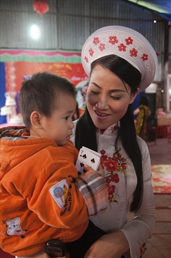 Relieved to have just been married, this bride looks adoringly at her relatives son. Reluctant to part with them, he clutches playing cards, a much loved traditional game across Vietnam and a great form of entertainment for the young during the long hours of a traditional wedding.