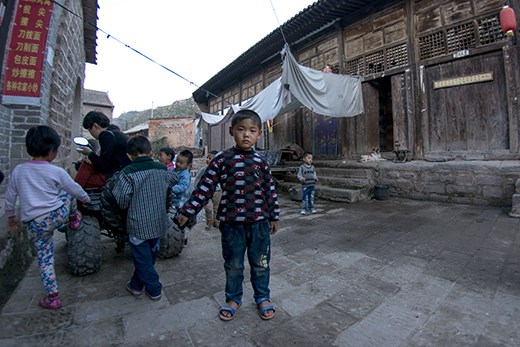 Children play around in the streets of Qikou, and a young boy, curious in the camera, step away to watch, while casually swinging his bag. Many people in these rural parts had never, or rarely, seen white people, so were fascinated in simply watching. 