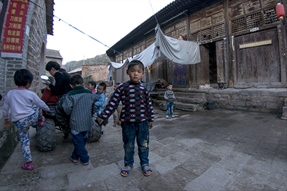 Children play around in the streets of Qikou, and a young boy, curious in the camera, step away to watch, while casually swinging his bag. Many people in these rural parts had never, or rarely, seen white people, so were fascinated in simply watching. 