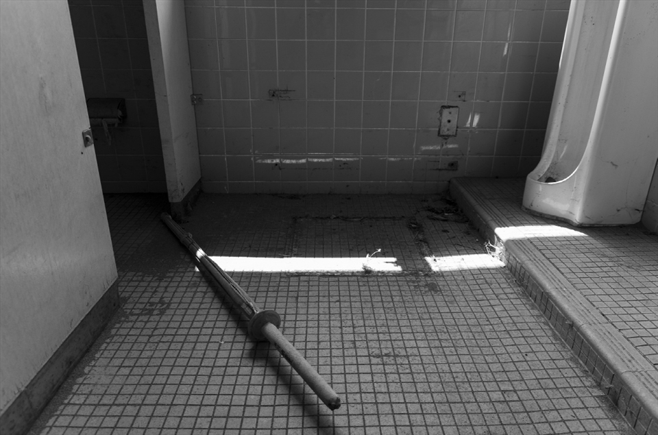 A kendo shinai rests on the floor of a boy's bathroom adjacent to the gymnasium.  Even though the buildings have been mostly cleaned out, items still remain as if to serve as an eerie reminder of the times once enjoyed here.