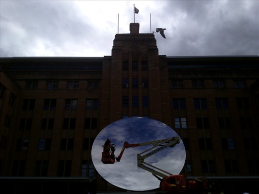 (4) Anish Kapoor is one of the most recognisable contemporary artists in the world today.  The Museum of Contemporary Art is 
featuring some of his works for the first time in the gallery. This photograph looks not only at Kapoor’s 
reflective work ‘The Sydney Eye’, it also looks at what an object is, in relation to other things. In this case, Kapoor’s circular work reflecting the sky around Sydney, is juxtaposed against the solid square matt mass of the museum itself.  It is fitting because Kapoor’s work looks at ideas of form, voids and content, and how an object can be and not be. I was particularly pleased to catch this 
moment of two workers shining the great reflective surface, preparing the work for the public gaze, themselves becoming a piece of art in my photograph. (This photo was taken with an iPhone)

