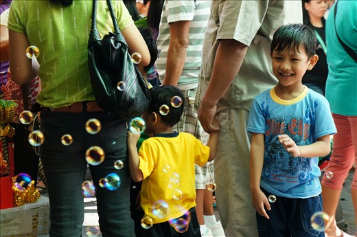 Unable to enter the packed church, some groups stay out on the street to hear mass. Merchants, oblivious to the ongoing mass, compete with the priest for their attention. A young boy turns away from his family and abandons listening to the sermon to gleefully play with the bubbles drifting temptingly in front of him.