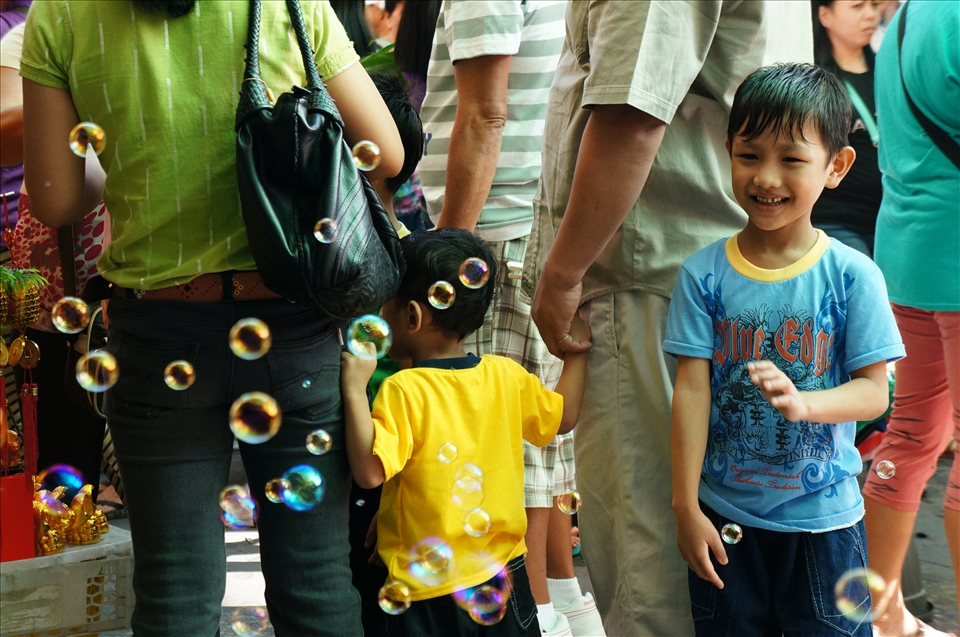 Unable to enter the packed church, some groups stay out on the street to hear mass. Merchants, oblivious to the ongoing mass, compete with the priest for their attention. A young boy turns away from his family and abandons listening to the sermon to gleefully play with the bubbles drifting temptingly in front of him.