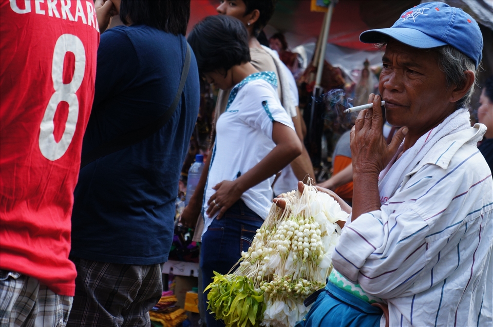 The smoke rising in languid curls into the air keeping him company, a Sampaguita (Jasmine flower) vendor battles fatigue with a stick of cigarrette while he waits at the back of the crowd for the chruchgoers to disperse. Most Filipinos treat their religious statues as human, which is believed to bring the owners luck: dressing them in elaborate clothing and sometimes offering food. Sampaguita necklaces are used to adorn religious statues' necks. 