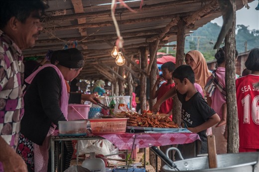 Markets along with food stalls are a very common sight in the entire country of Thailand. This image was taken on the island Koh Lanta, which has a very diverse cultural mix. Thai-Muslim, Thai-Chinese and Sea Gypsies all living together, this can make food shopping very interesting. You can never be short of a bite to eat, when the early markets are finished, there is always the option to go find the evening market. The light blub hanging over as the local boy makes his selection is powered by the electric power cables that run above the roadside.  