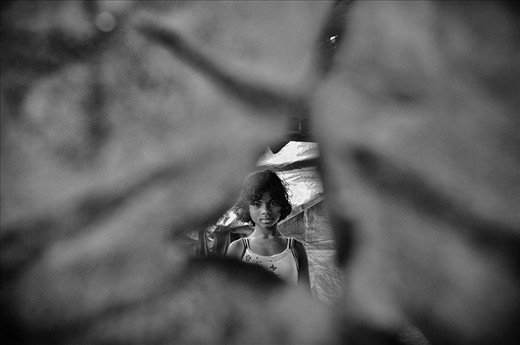 Moromi looks out of the torn shards of a tarpaulin she currently lives in. She lived in a fine house amidst towering areca nut trees in her quaint village, Dotma. Today, she lives in a relief camp, just three hundred meters away from the site of what was once home. Eight months back, her district Kokrajhar become the epicenter of riots tossing her life into uncertainty of a dark gloom.
