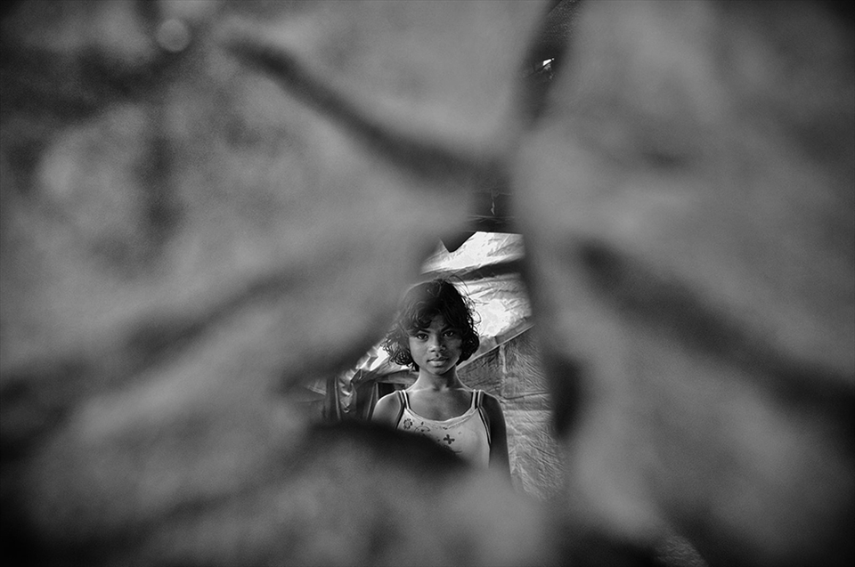 Moromi looks out of the torn shards of a tarpaulin she currently lives in. She lived in a fine house amidst towering areca nut trees in her quaint village, Dotma. Today, she lives in a relief camp, just three hundred meters away from the site of what was once home. Eight months back, her district Kokrajhar become the epicenter of riots tossing her life into uncertainty of a dark gloom.