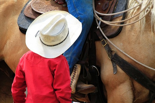 A young boy looks up at an older cowboy, his father, sitting on his horse. This young boy is also able to rope calves and throw a lasso, but is too young to compete just yet.