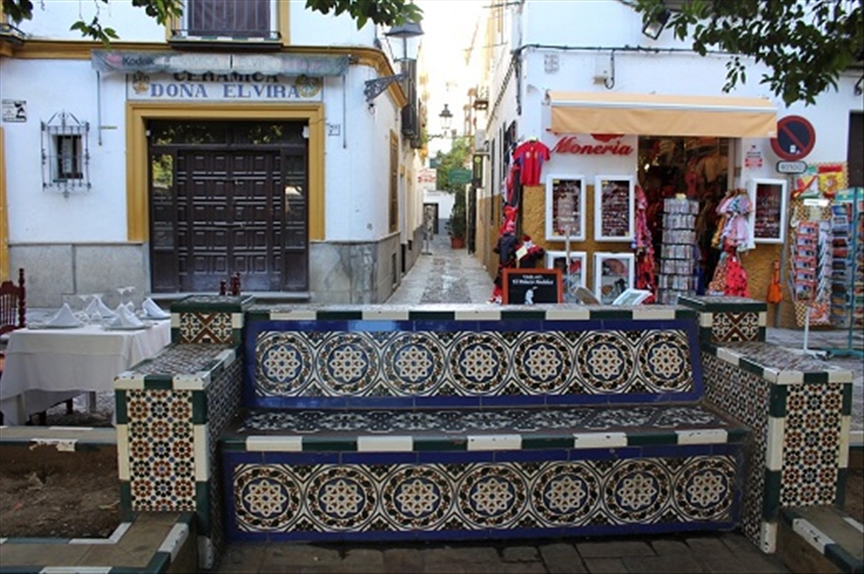 This is a very nice mosaic bench in Seville, Spain. You can also  find a narrow  street ( callejuelas  in Spanish word) in the  background (middle) of this image.   At  the right side is a souvenir store,  where you can buy  souvenirs of all kinds from postcards to  travel guides, fridge magnets, shirts, etc. At the left side a ceramic  shop. 