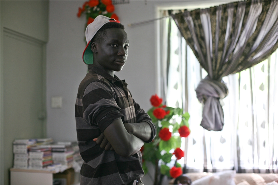 Joel Okumu, standing confidently in living area of family home.