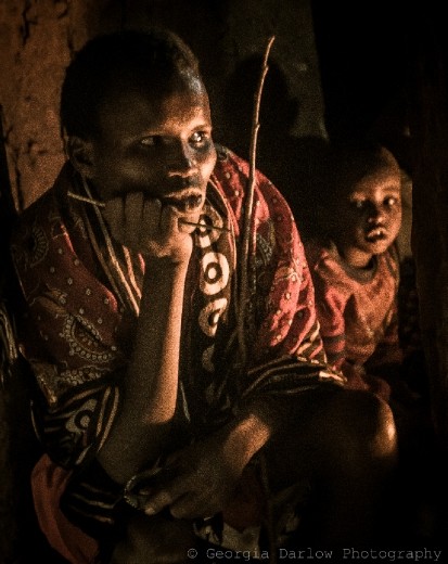 Members of a Maasai family sit inside a house, lit with only the light of the fire. 