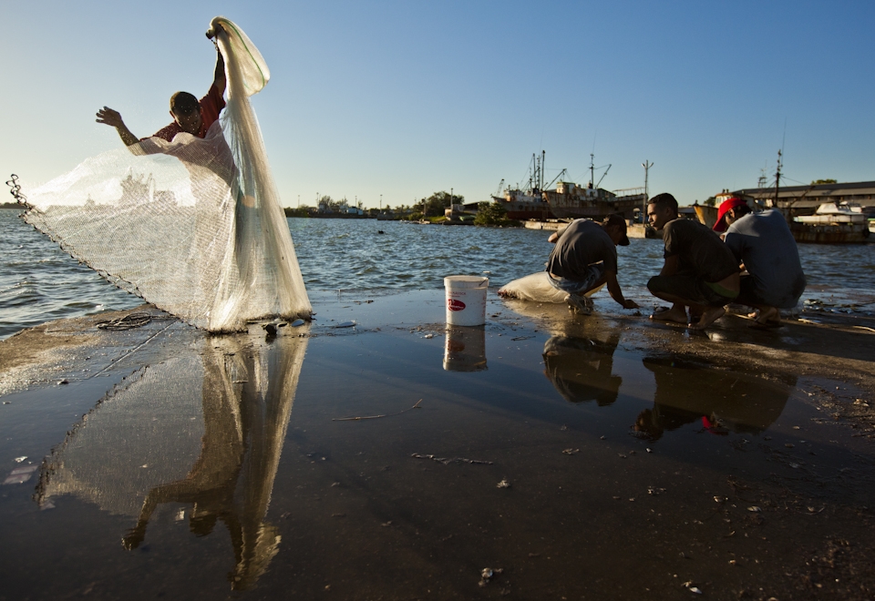 A young fisherman In Cienfuegos, Cuba watches an older man unfurl his net while he cuts bait. Fishermen come to the central pier just before dusk every night to catch the white fish that are then sold to local restaurants and fish mongers.
