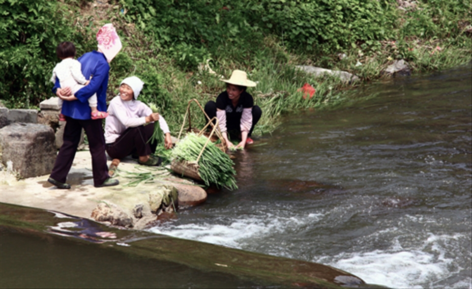 A grandmother carrying her grandchild on the back, chats to two farmer women who are washing the crops in the village river.

The majority of traditional Dong villages are settled along rivers. The water is their centre of life.