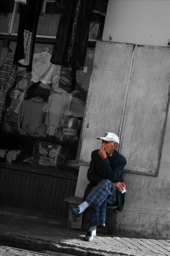 A quiet day at work. A man is sittin outside his shop looking at all the people passing. 