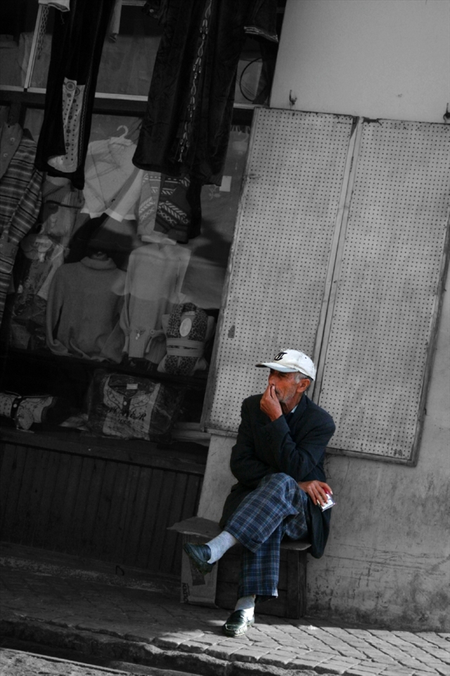 A quiet day at work. A man is sittin outside his shop looking at all the people passing. 