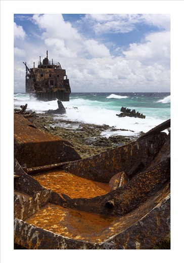 A shipwreck, torn apart and strew about a beach is battered by waves. The sea is rough on this side of the small spit of land. The sea washes over the ships rusty remains.