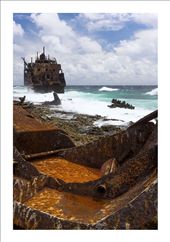 A shipwreck, torn apart and strew about a beach is battered by waves. The sea is rough on this side of the small spit of land. The sea washes over the ships rusty remains.: by hoffeman, Views[899]